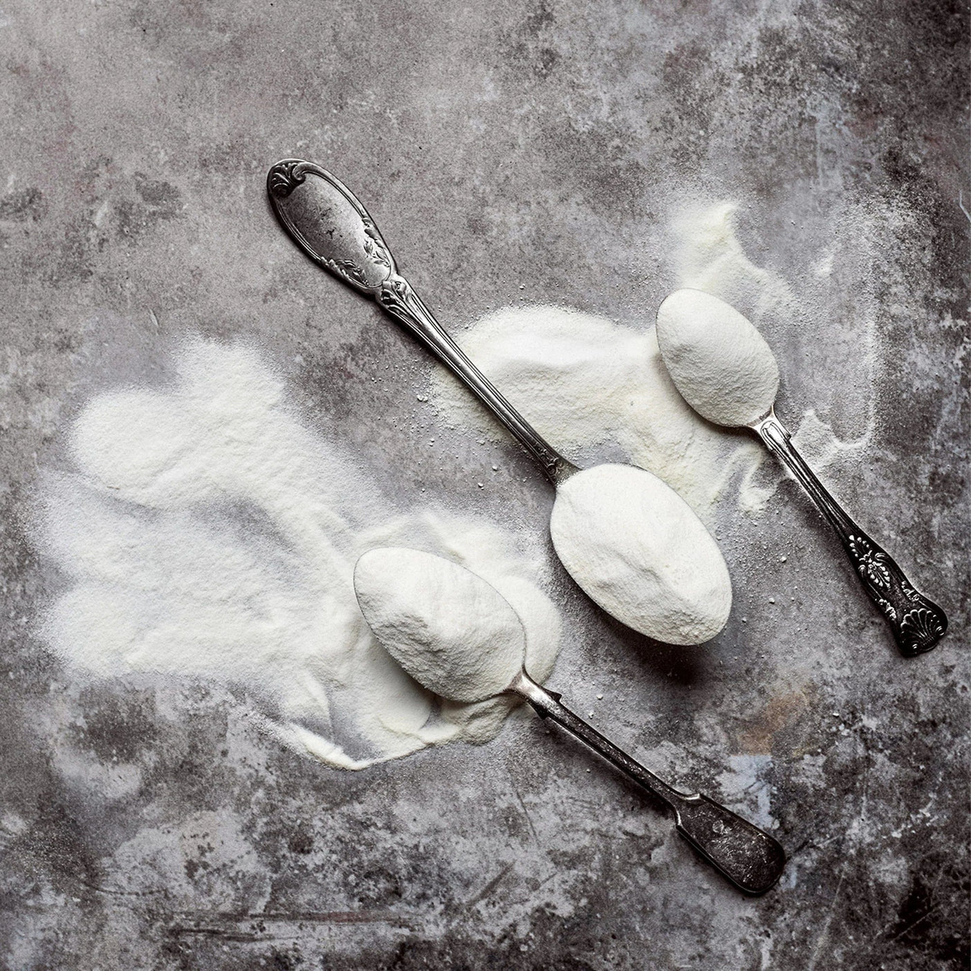Two spoons full of collagen powder on a textured grey surface.