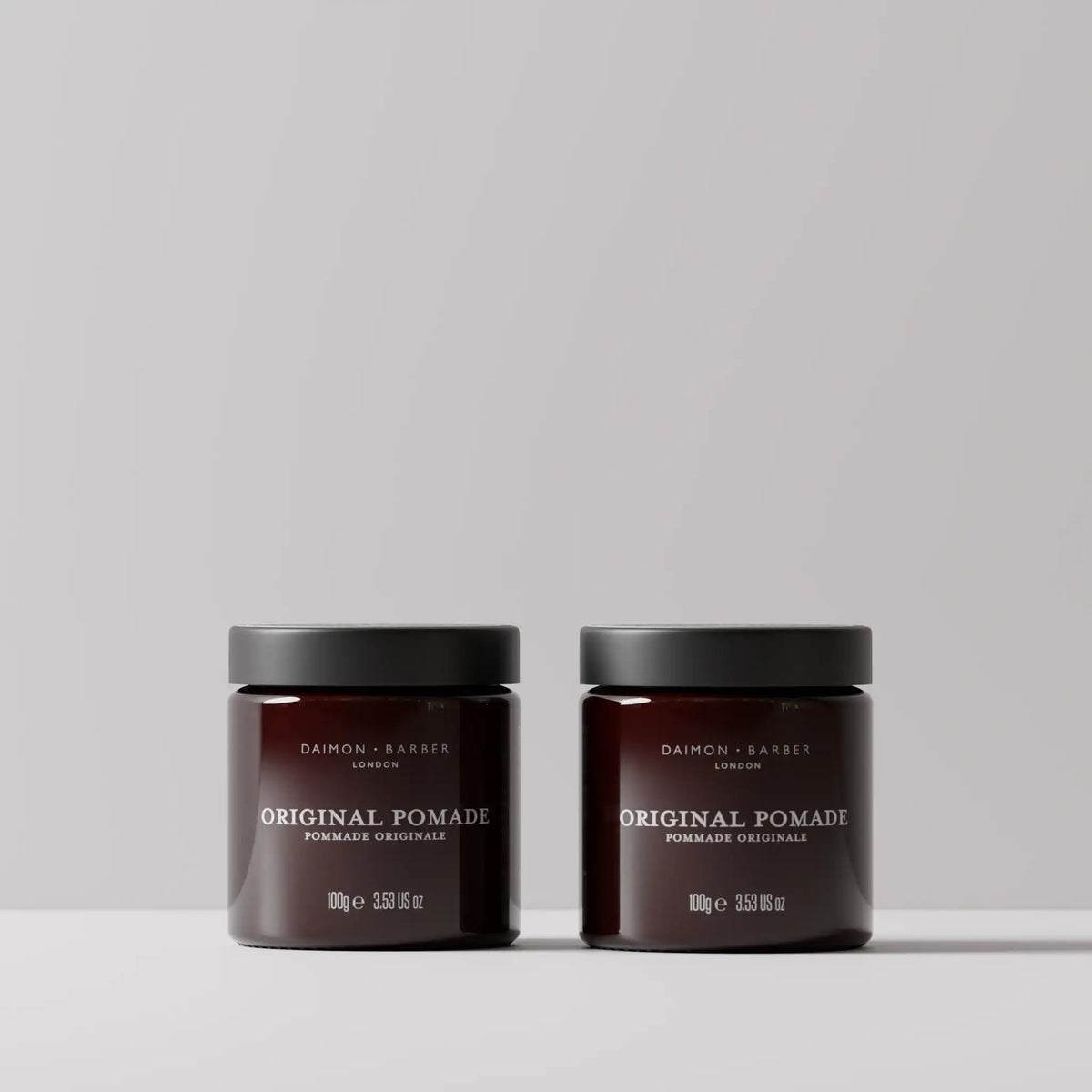 Two claret glass hair styling product jars labeled Daimon Barber Texture Clay, shown side by side on a light gray surface against a neutral gray background.