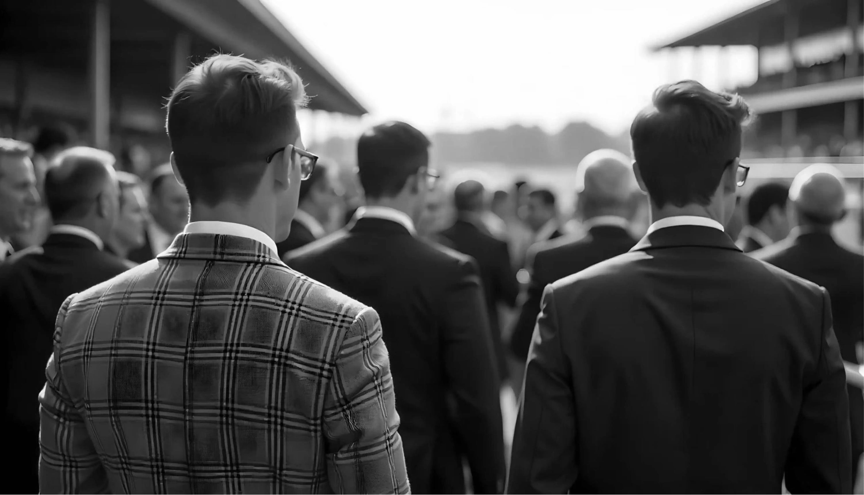 Men in suits looking onto horse racing course in the stands in black and white