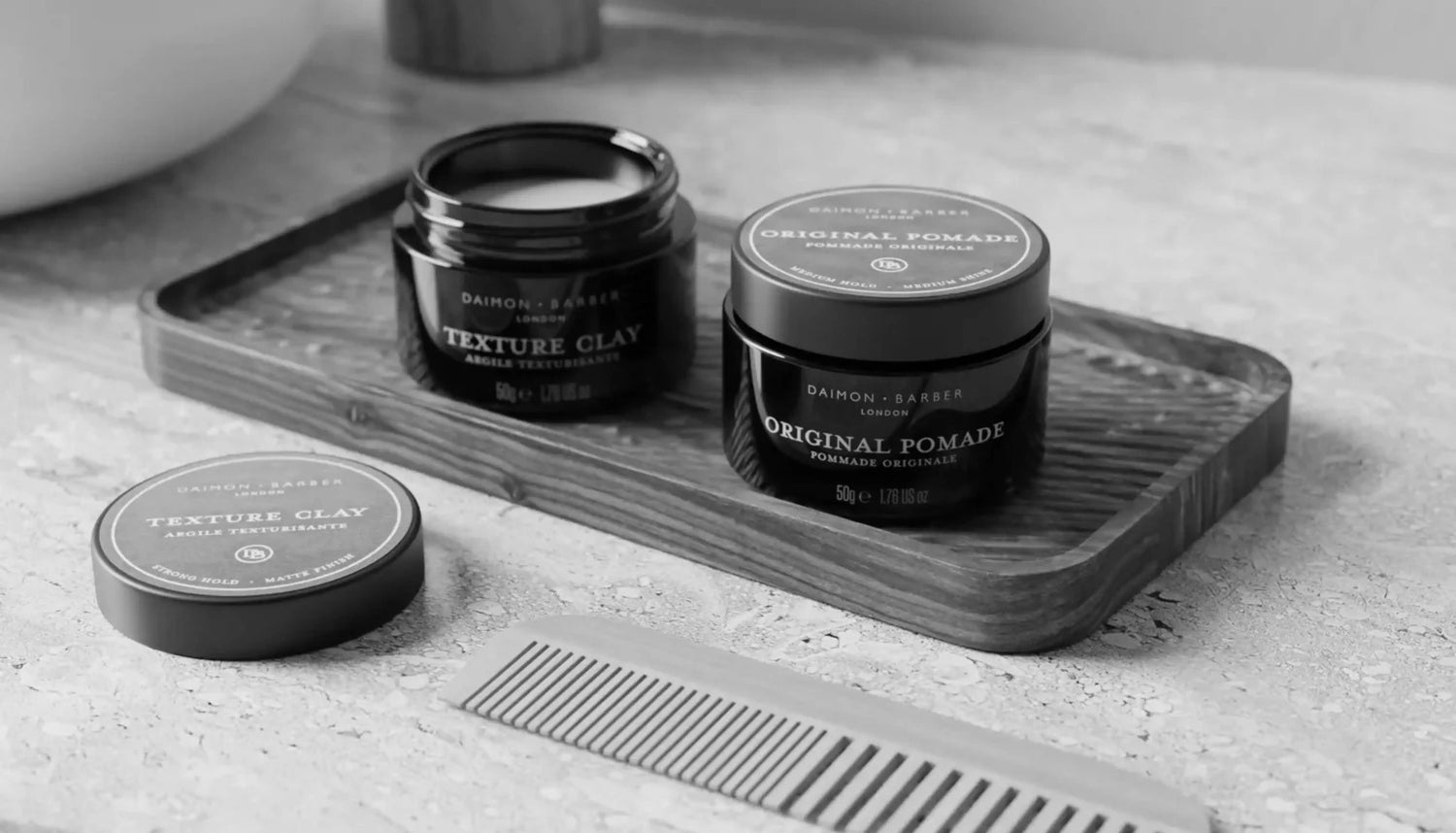 Open pot of Texture Clay and Original Pomade next to a bathroom sink with a comb for men’s hair styling in black and white