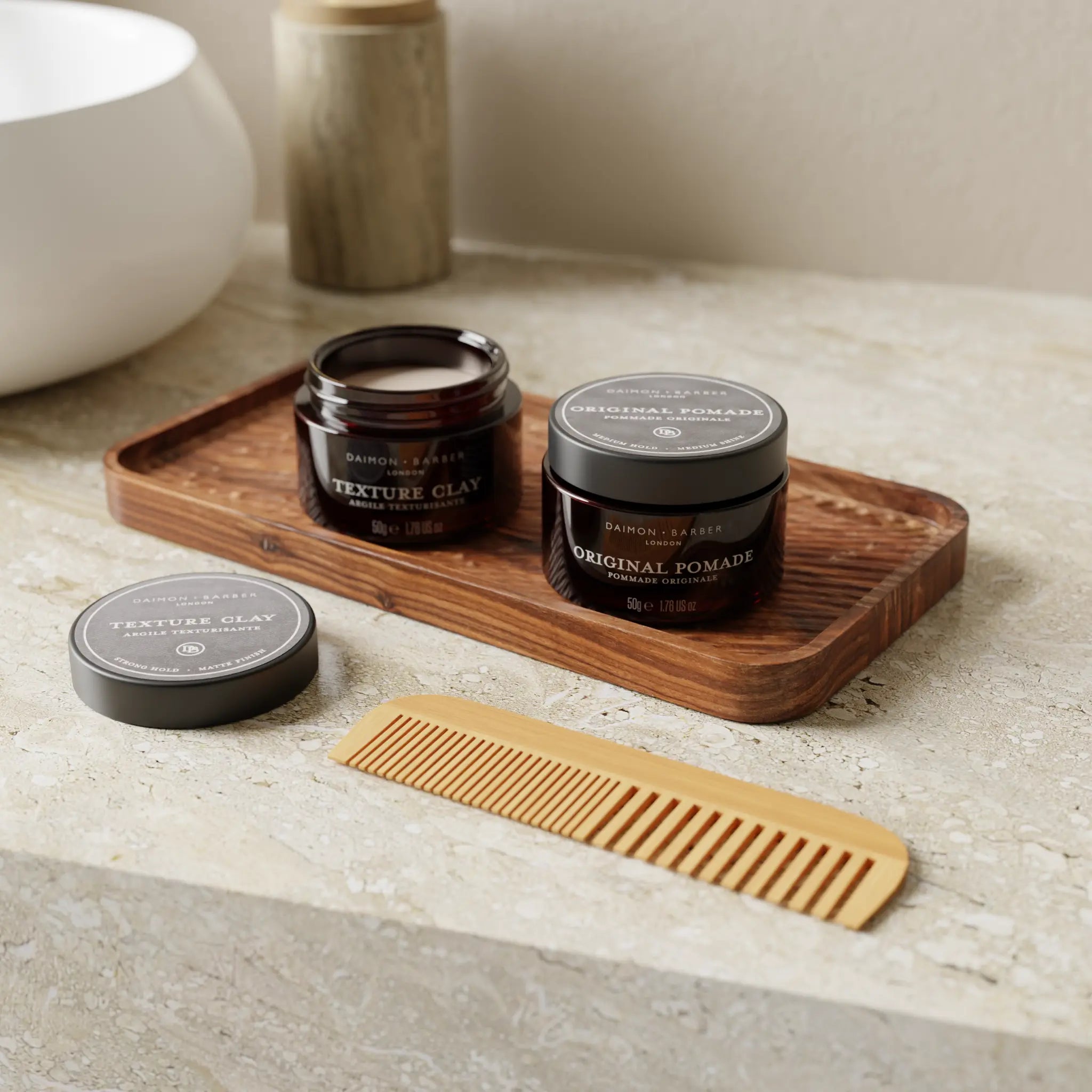 Open pot of Texture Clay and Original Pomade next to a bathroom sink with a comb for men’s hair styling.