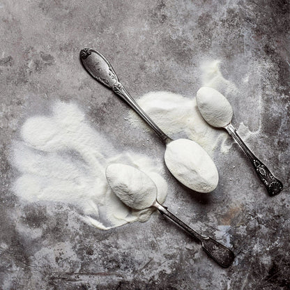 Two spoons full of collagen powder on a textured grey surface.
