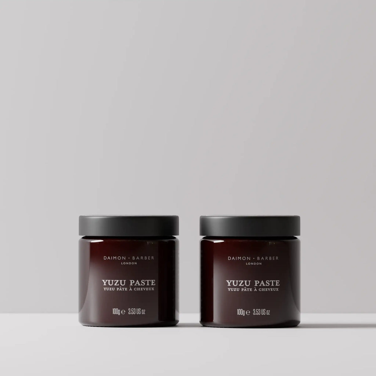 Two claret glass jars of Daimon Barber Yuzu Paste with black lids, displayed side-by-side on a light gray surface against a neutral light gray background.